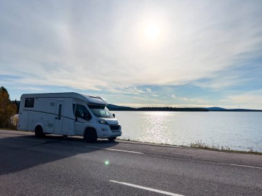 Güneşli bir günde gölün kenarına park edilmiş beyaz bir karavan manzarası. Yolculuk ve tatil için ideal.