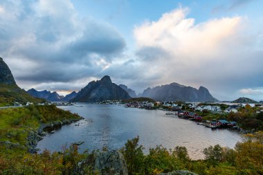 Norveç 'in kuzeyindeki Reine, Lofoten Adaları' nda sonbahar günbatımı, dramatik dağlar, sakin sular ve kıyı boyunca geleneksel evler..