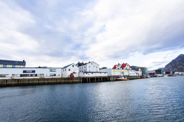 A scenic view of Henningsvaer in Lofoten Islands, Norway, showcasing traditional buildings along the harbor with mountains in the background. clipart