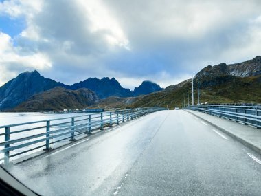 Norveç 'te bir fiyortta manzaralı bir yolda giden beyaz bir karavan. Arka planda dağlar ve bulutlu bir gökyüzü var.