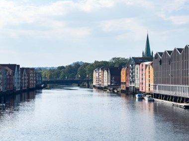 Traditional colorful wooden houses lining the nidelva river in trondheim, norway, creating a picturesque reflection on a sunny day clipart