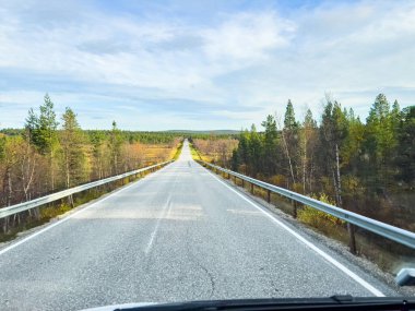 Terkedilmiş otoyolda, açık mavi gökyüzü altında, tüylü bulutlar altında, engin sonbahar manzarası boyunca giden araç gölgesini düşürüyor.