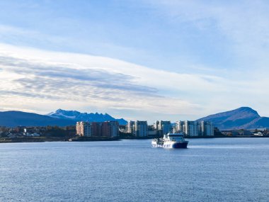 Yolcu feribotu, karla kaplı dağları ve bulutlu gökyüzü olan modern bir şehrin sahil şeridine doğru gidiyor.