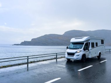 Yağmurlu bir sonbahar gününde Norveç kıyıları boyunca manzaralı bir yolda giden beyaz bir karavan.