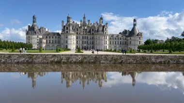Bahçelerde yürüyen turistlerle dolu Chambord Şatosu mavi gökyüzünün altındaki suya yansıyor.