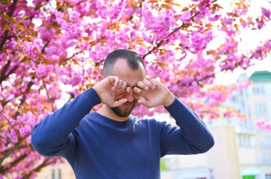 A man is standing near a tree with flowers but appears to be suffering from allergies. He looks uncomfortable, rubbing his nose and eyes, and perhaps considering taking allergy medication. clipart