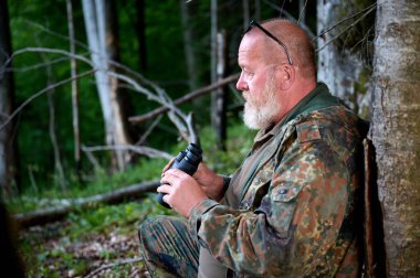 Avlanırken elinde dürbün ve omzunda bir tüfek tutan gri sakallı kıdemli avcı, ormanda yürüyor, dürbünden bakıyor ve doğayı gözlemliyor. Avlanma ve seyahat kavramı