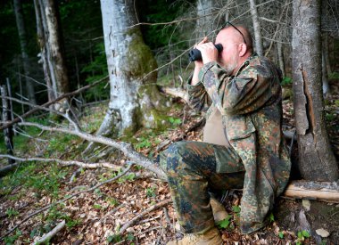 Ellerinde dürbün ve omzunda tüfekle gezen bir erkek avcı ormanda yürüyor, dürbünden bakıyor ve doğayı gözlemliyor.