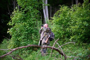 Ellerinde dürbün ve omzunda tüfekle gezen bir erkek avcı ormanda yürüyor, dürbünden bakıyor ve doğayı gözlemliyor.