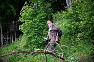 Ellerinde dürbün ve omzunda tüfekle gezen bir erkek avcı ormanda yürüyor, dürbünden bakıyor ve doğayı gözlemliyor.