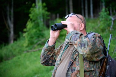 Ellerinde dürbün ve omzunda tüfekle gezen bir erkek avcı ormanda yürüyor, dürbünden bakıyor ve doğayı gözlemliyor.