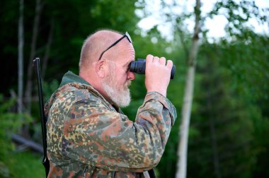 Ellerinde dürbün ve omzunda tüfekle gezen bir erkek avcı ormanda yürüyor, dürbünden bakıyor ve doğayı gözlemliyor.