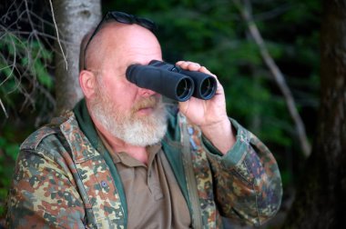 Ellerinde dürbün ve omzunda tüfekle gezen bir erkek avcı ormanda yürüyor, dürbünden bakıyor ve doğayı gözlemliyor.