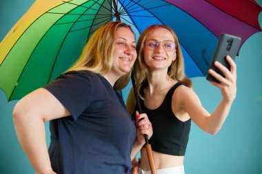 Renkli bir şemsiye altında sarılan iki mutlu lezbiyen kız. Çekici kadın desteği, lezbiyen çift kucaklaşması. LGBTQ bayrak renginde şemsiye altında selfie çeken iki bayan arkadaş..