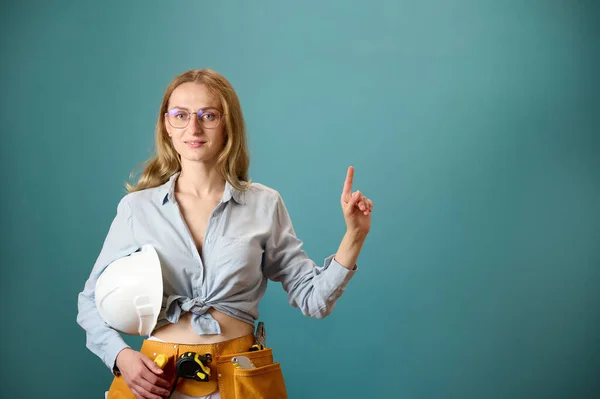 Young female worker with tool belt holding an instrument in hands isolated on background. Pretty caucasian female with tools planning new project. Portrait in studio. Copy space.