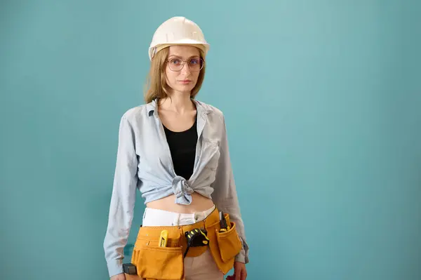 Young female worker with tool belt holding an instrument in hands isolated on background. Pretty caucasian female with tools planning new project. Portrait in studio. Copy space.