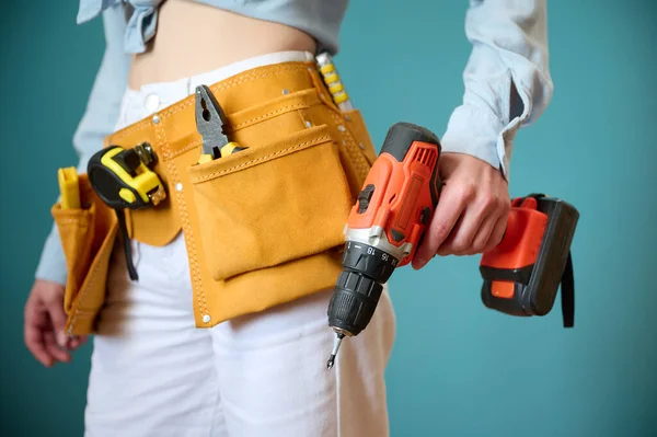 Young female worker with tool belt holding an electric screwdriver in hands isolated on background. Pretty caucasian female with tools planning new project. Portrait in studio.