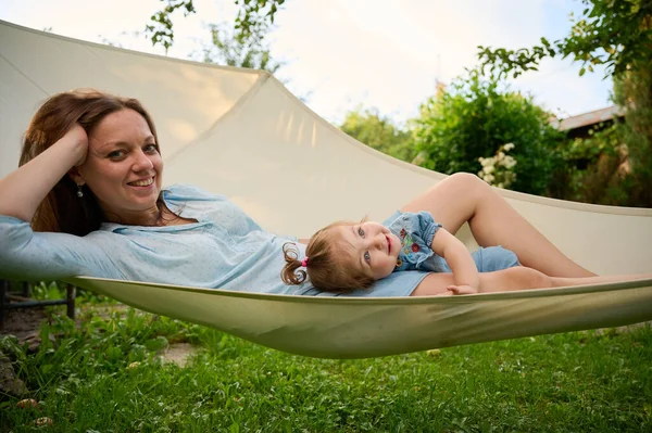 Mommy and her doughter playing in the garden, lying in hammock. Lovely family posing outdoors. Happy little girl in summer scenery. Sweet small kid outdoors.