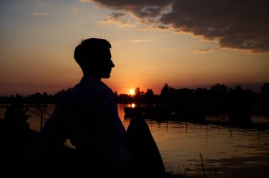 Çimenlerde oturmuş günbatımının tadını çıkaran kısa saçlı genç kız silueti. Günlük giysiler içinde güzel bir kadın açık havada hayatın tadını çıkarıyor. Gölet.