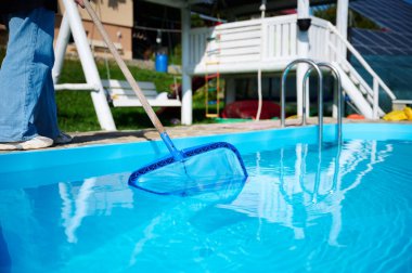 Genç kadın yüzme havuzu temizliyor. Güneşli yaz günlerinde havuzu yapraklardan temizleyen personel. Otel çalışanı havuzu temizliyor. Yüzme havuzu servisi. Ağ ile arındırma. Kapat.