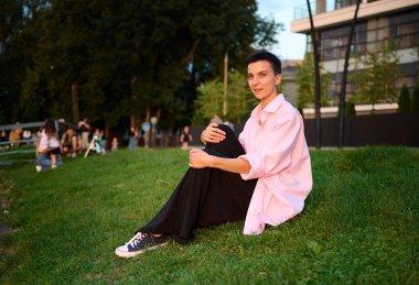 Genç, kısa saçlı bir kız yeşil çimenlerde oturmuş günbatımının tadını çıkarıyor. Günlük giysiler içinde güzel bir kadın açık havada hayatın tadını çıkarıyor. Kentsel alanda yeşil bölge.