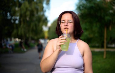 Güneşli bir günde limonata içen güzel bir kız. Genç bir kadın güneş ışığı yüzünden gözlerini kısıyor. Olumlu kız dışarıda içiyor ve dinleniyor..