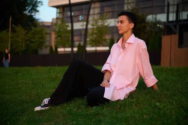 Young short-haired girl is sitting on a green lawn enjoying the sunset.Pretty female in casual clothes enjoying life outdoor. Green zone in urban space.