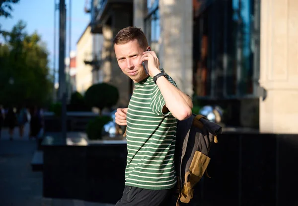 Young sporty man in a striped T-shirt walking on the street with a smartphone in hand and a trendy hipster\'s backpack slung over his shoulder, enjoying a leisurely urban outing.