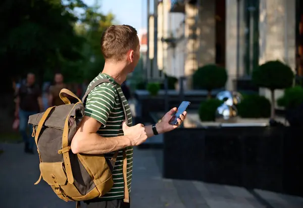 Çizgili tişörtlü genç bir adam elinde akıllı bir telefonla caddede yürüyor ve modaya uygun bir hipsterin sırt çantası omzunda gezintiden keyif alıyor..