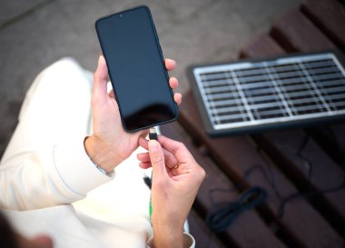 Güzel bir kadın bankta oturuyor ve telefonunu taşınabilir güneş paneliyle şarj ediyor. Mobil cihazlar için güneş pili. Sürdürülebilir yaşam tarzı ve yeşil yenilenebilir enerji kavramı. Ekin görünümü