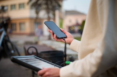 Güzel bir kadın bankta oturuyor ve telefonunu taşınabilir güneş paneliyle şarj ediyor. Mobil cihazlar için güneş pili. Sürdürülebilir yaşam tarzı ve yeşil yenilenebilir enerji kavramı. Ekin görünümü