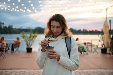 Sahilde yürüyen, çay içen, selfie çeken ve telefonda sosyal ağlara göz atan güzel bir kadının portresi. Sahilde güzel bir gün batımı ve bir sahil kafede güzel bir yapay ışıklandırma.