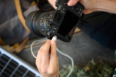 Kamerasını şarj etmek için güneş paneli kullanan gezgin fotoğrafçı. Genç gezgin kamerasının şarj olmasını bekliyor. Pozitif erkek uzun bir yürüyüşten sonra dinleniyor..