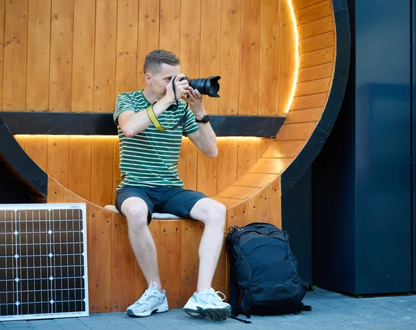 Excited photographer looks at the photos he just took. Young photographer sitting and checking result of photosession. Handsome male sitting on round bench with solar panel on background.