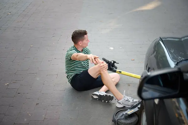 Dikkatsiz adam elektrikli scooter 'dan düştü. Genç erkek, araçtan düştükten sonra sırtüstü yatıyor. İlham perisi bir arabayla çarpıştı ve dizlerini incitti..
