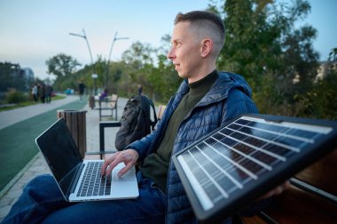 Kendine güvenen iş adamı bankta oturuyor ve dizüstü bilgisayarını taşınabilir güneş paneliyle şarj ediyor. Yenilenebilir enerji kavramı. Yeşil bağımsız enerji. Yan görünüm