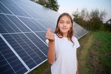 Güneş panellerinin yanında kalıp kalp sembolü gösteren güzel bir genç kız. Güneş enerjisi kavramı. Yenilenebilir enerji.