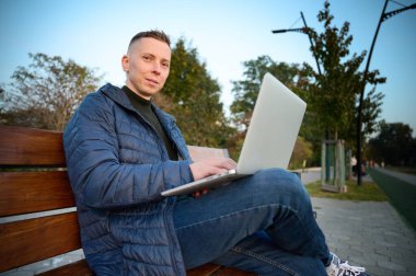 Kendine güvenen iş adamı gün batımında bankta oturuyor ve dizüstü bilgisayarı üzerinde çalışıyor. Parkta otururken bilgisayardan uzaktan çalışan adamın portresi.