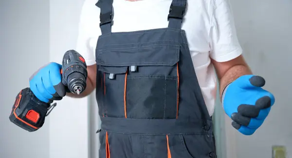 Construction worker holding electric screwdriver in hand at construction site. Accessories for assembling, install furniture, repair home. Man dressed in work attire and helmet and protective glasses.