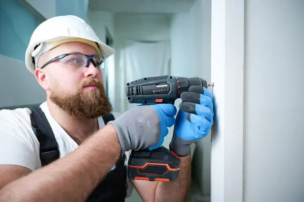 Construction worker holding electric cordless screwdriver in hand. Accessories for install furniture, repair home. Man worker dressed in work attire, helmet and protective glasses. Home renovation