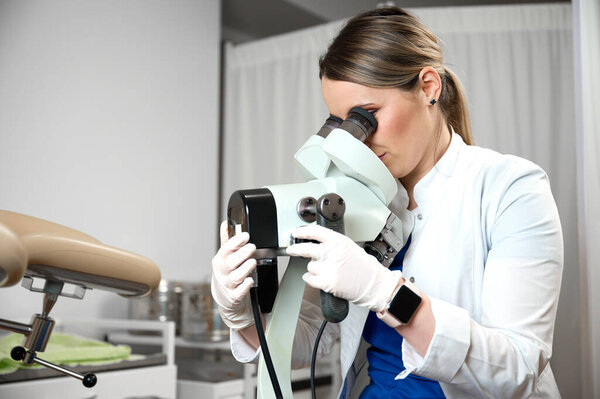 Pretty female doctor in medical sterile gloves preparing for colposcopy. Gynecologist woman sitting near a colposcope in her gynecological office. Women's health. Early detection of cancer.