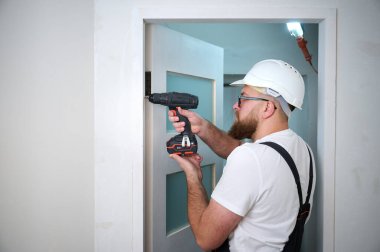 Kapı monte eden tornavidayla inşaat işçisi. Montaj için aksesuarlar, mobilya monte etmek, evi onarmak için. İş kıyafeti, kask ve koruyucu gözlük takmış bir adam. Ev yenileme konsepti