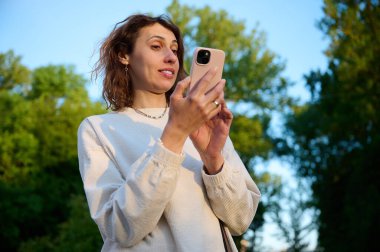 Parkta yürüyen ve telefonla konuşan, sıradan giyinmiş, şaşırmış genç bir kadının portresi. Parkta telefonla konuşan genç şokta bir kadın..