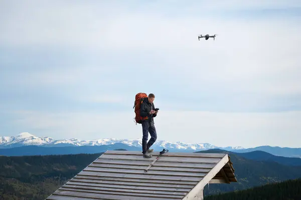 stock image Fully equipped hiker staying on the top of the roof of the wooden cabin in the mountains. Drone operator holding remote controller. Traveler controlling drone flying over mountains with remote control