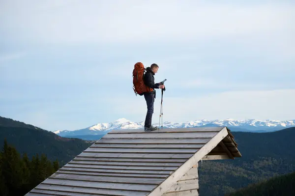 stock image Fully equipped hiker with backpack and trekking poles staying on the top of the roof of the wooden cabin in the mountains. Traveler standing gazing out at the breathtaking landscape before them.