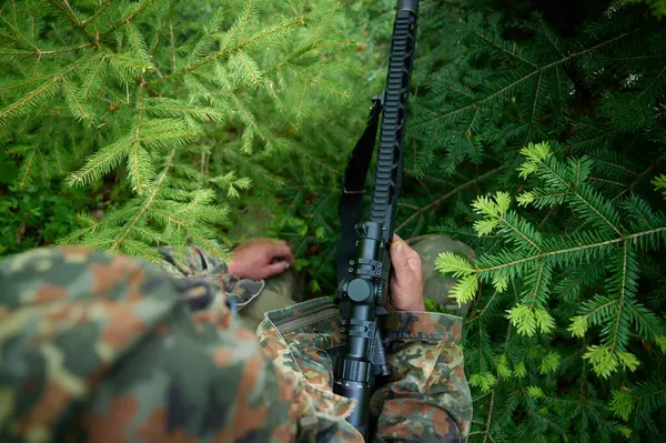 stock image Hunter with rifle wearing camouflage suit sitting in ambush. Camouflaged sniper in the forest in ambush. Military man hide in the woods with rifle.