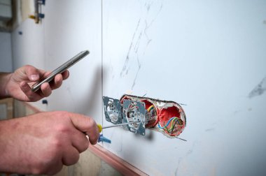 Profesyonel elektrikçi yeni bir evde tornavida kullanarak duvara elektrik prizi takıyor. Evde elektrik tamiratı var. Elektrikçi prizi elektrikli tornavidayla bağlıyor.