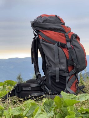 Turist macerası sırt çantası, sırt çantasıyla dağlara karşı tepenin üstünde duruyor. Çimlerin üzerinde duran turist yürüyüşü çantasıyla doluydu. Kamp ve yürüyüş ekipmanları