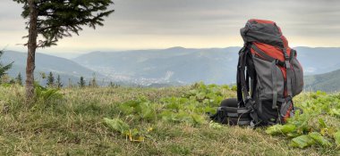 Turist macerası sırt çantası, sırt çantasıyla dağlara karşı tepenin üstünde duruyor. Çimlerin üzerinde duran turist yürüyüşü çantasıyla doluydu. Kamp ve yürüyüş ekipmanları