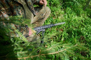 Hunter with rifle wearing camouflage suit sitting in ambush. Camouflaged sniper in the forest in ambush. Military man hide in the woods with rifle. clipart